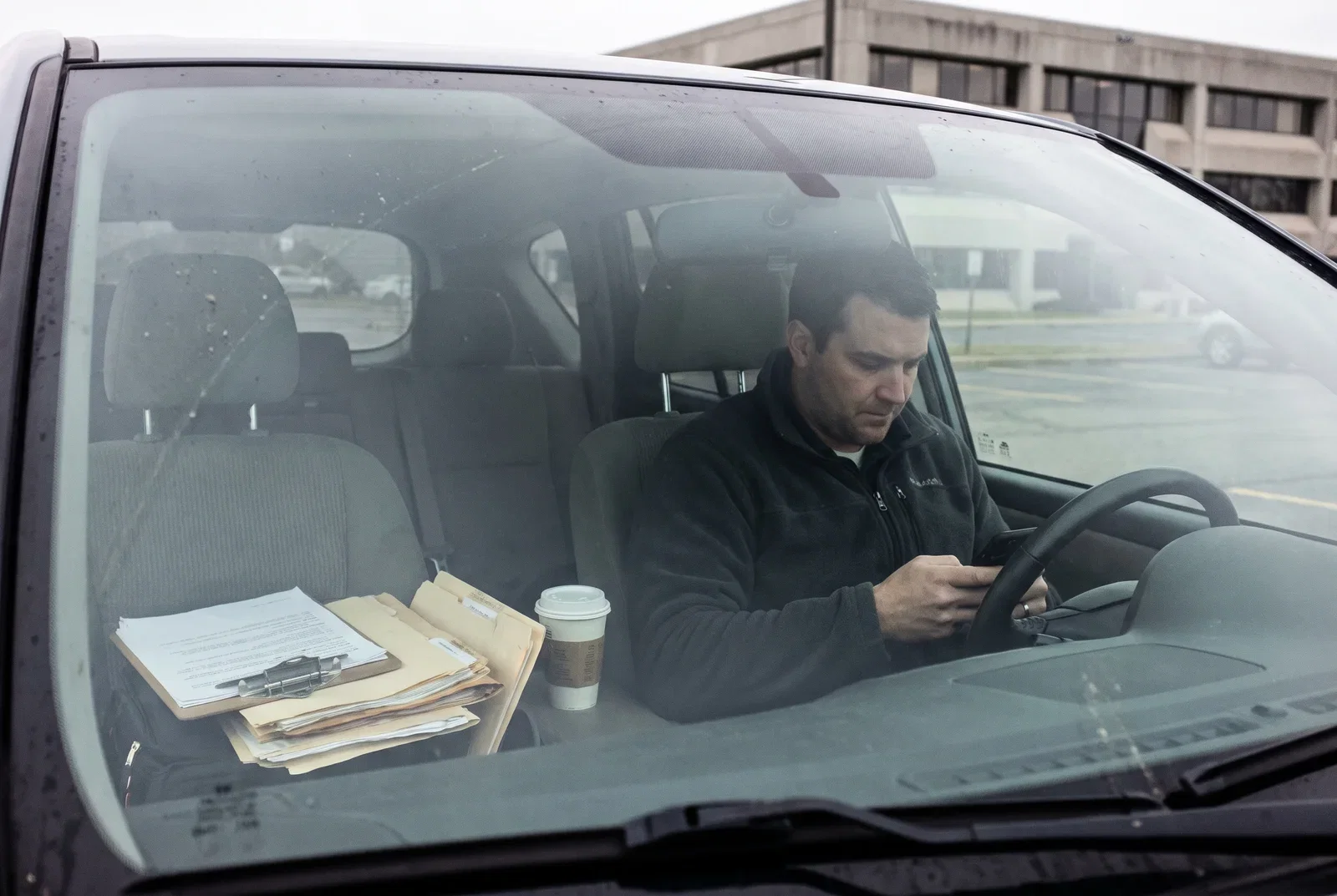 Ein Betriebsinhaber sitzt im Auto auf dem Firmenparkplatz und sucht auf dem Smartphone seine eigene Leistung, auf dem Beifahrersitz liegen Unterlagen und ein Klemmbrett